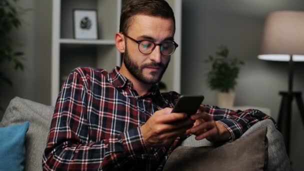 Gros plan d'un jeune homme heureux assis sur le canapé tout en envoyant des SMS sur le smartphone et en regardant la caméra, souriant joyeusement sur le fond du salon le soir. Plan intérieur . — Video