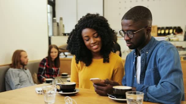 Amigos afro-americanos alegres aproveitando seu tempo livre no café da cidade, bebendo café e usando um telefone celular enquanto conversam um com o outro durante o dia. Tiro interior . — Vídeo de Stock