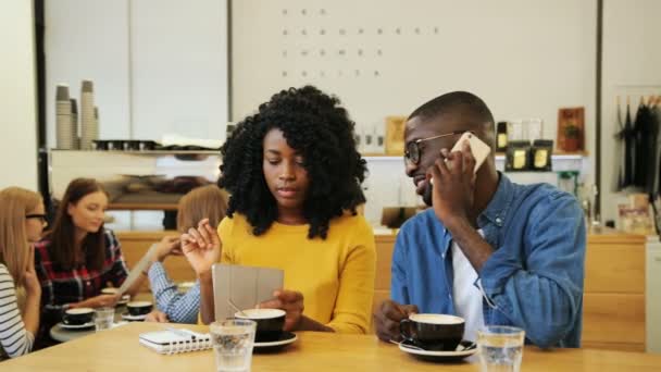 Glada Afro-American vänner njuta av sin fritid i staden coffee shop, dricka kaffe och arbetar på tablett enheten och talar på mobilen och chattar med varandra på café — Stockvideo