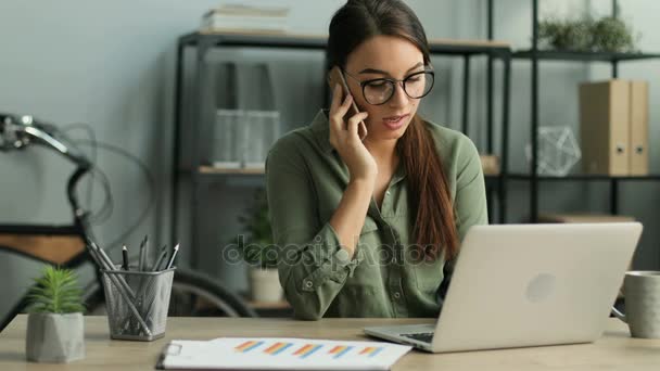 Attraktiva affärskvinna talar på mobilen medan du skriver på laptop tangentbord i thr office. Kvinnan håller telefonen mellan huvudet och axeln. närbild — Stockvideo