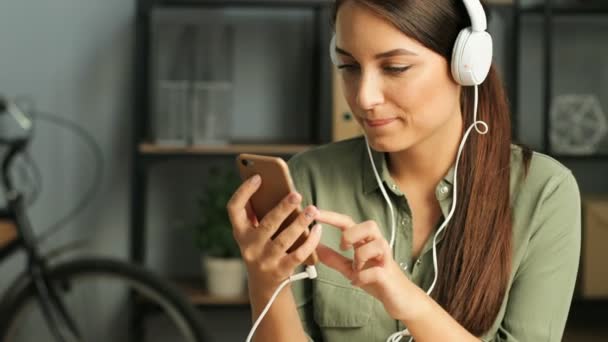 Attraktive Frau, die in der Pause im Büro mit ihrem Smartphone Musik in weißen Kopfhörern hört. Nahaufnahme — Stockvideo