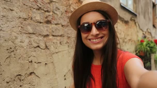 Portret shot van mooie jonge vrouw in hoed en zonnebril zwaaien naar de camera en glimlachen. Vrouw videochat buitenshuis in de oude straat van de stad. Close-up — Stockvideo