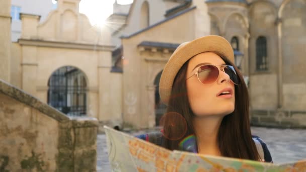 Hermosa joven con sombrero y gafas de sol utilizando el mapa de papel turístico y mirando a su alrededor en la hermosa calle vieja. Mujer impresionada con la arquitectura. de cerca — Vídeo de stock