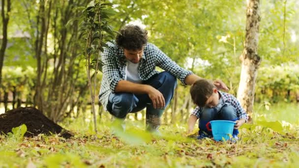 Een portret van een kleine jongen en zijn papa tuinieren. Jongen zet bodem in zijn emmer. Papa kranen hem op zijn schouder. Onscherpe achtergrond — Stockvideo