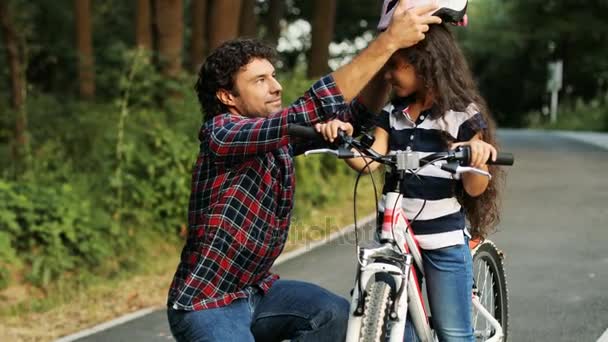 Retrato de una niña y su padre cerca de la bicicleta. Papá lleva un casco en la cabeza de las chicas. Se miran el uno al otro, entonces - en la cámara. Papá abrazando a su hija. Sonriendo. Fondo borroso — Vídeo de stock