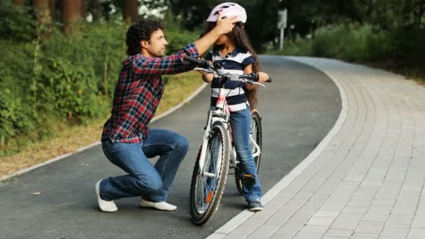 Primer plano. Retrato de una chica bonita y su padre cerca de la bicicleta. Papá lleva un casco en la cabeza de las chicas. Sonriendo. Fondo borroso — Vídeo de stock