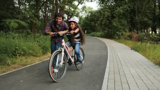 Primer plano. Retrato de una niña. Su padre le está enseñando a andar en bicicleta. Él la deja ir. Riendo. Cámara móvil. Fondo borroso — Vídeos de Stock