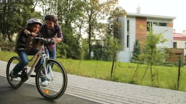 Nahaufnahme. Porträt eines kleinen Mädchens. Ihr Vater bringt ihr das Fahrradfahren bei. Er lässt sie gehen. glückliches Mädchen, das in die Kamera blickt. bewegte Kamera. verschwommener Hintergrund — Stockvideo