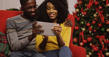 Noel Noel ağacı arkalarında, Oda tutarak ve tablet cihaz ekran üzerinde seyir dekore edilmiş rahat içinde kırmızı kanepede oturan aşık genç Afrikalı-Amerikalı çift. Kapalı