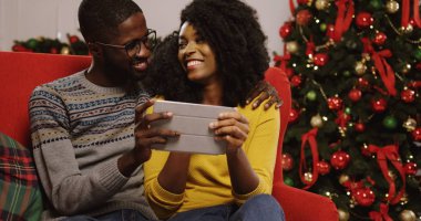 Noel Noel ağacı arkalarında, Oda tutarak ve tablet cihaz ekran üzerinde seyir dekore edilmiş rahat içinde kırmızı kanepede oturan aşık genç Afrikalı-Amerikalı çift. Kapalı