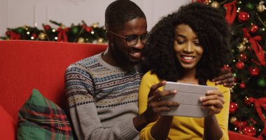 Noel Noel ağacı arkalarında, Oda tutarak ve tablet cihaz ekran üzerinde seyir dekore edilmiş rahat içinde kırmızı kanepede oturan aşık genç Afrikalı-Amerikalı çift. Kapalı