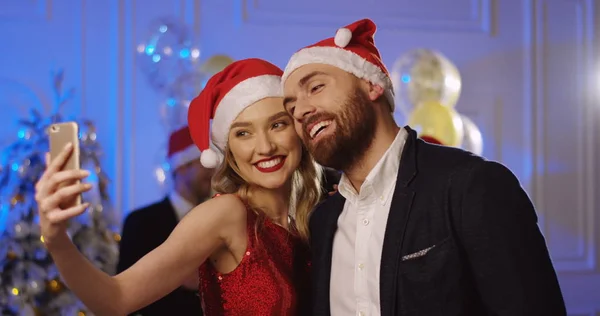 Portrait shot of young attractive couple in santa clause hats making funny selfies on a smart phone at the Christmas party. Dancing people background. Indoors