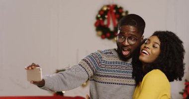 Komik selfies oturma odası Noel atmosferini bir akıllı telefon alarak aşık genç mutlu Afrika kökenli Amerikalı çift. Noel ağacı arka planda. Kapalı