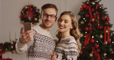 Portre gülümseyen selfies bir akıllı telefon yapma ve zafer gösterilen aşık genç mutlu çiftin güzel bir şekilde dekore edilmiş oturma odasında imzalar. Noel ağacı arka plan.