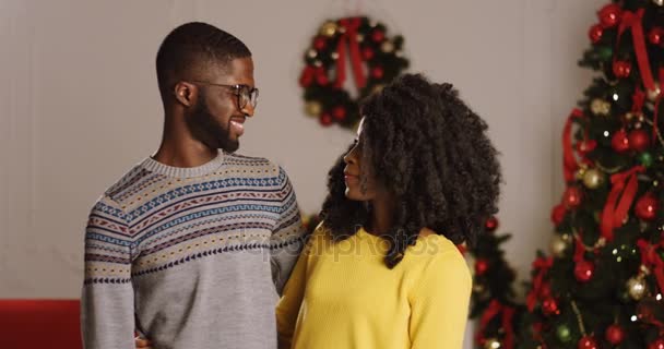 Молодая счастливая афроамериканская пара смотрит друг на друга и чем обнимает и улыбается в камеру на рождественской елке с подсветкой. В помещении . — стоковое видео