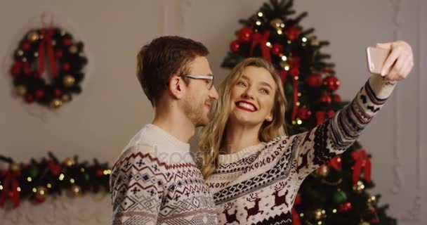 Joven pareja feliz en el amor haciendo selfies divertidos en un teléfono inteligente y besándose en el ambiente de Navidad de la sala de estar. Árbol de Navidad en el fondo. De interior — Vídeo de stock