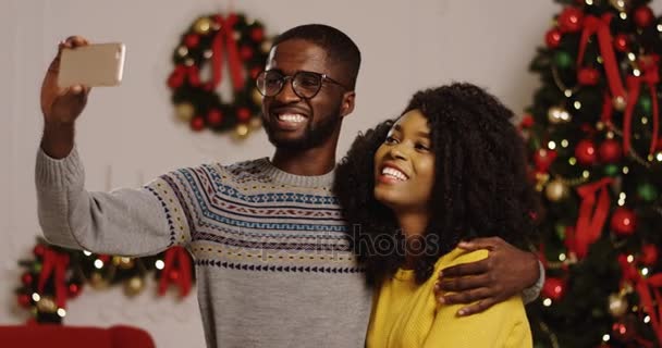 Portrait tourné de jeunes souriants et heureux couple afro-américain ayant vidéochat sur un téléphone intelligent dans le salon décoré confortable. Arbre de Noël avec lumières et rubans rouges sur le fond — Video