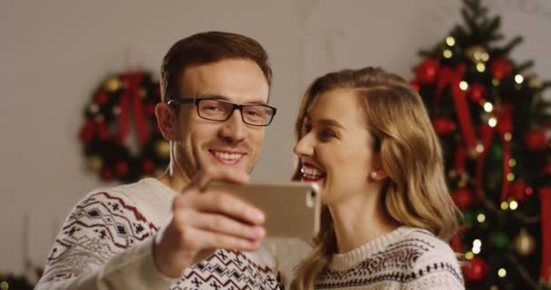 Nahaufnahme eines jungen lächelnden und glücklichen Paares, das im gemütlich dekorierten Wohnzimmer auf einem Smartphone einen Videochat führt. Weihnachtsbaum Hintergrund. Innenraum. Porträtaufnahme. — Stockvideo