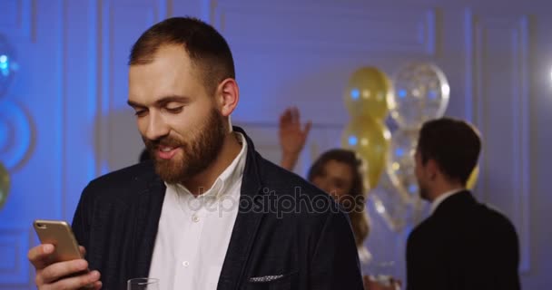 Jovem atraente gravando em seu telefone inteligente e bebendo champanhe na festa. Dançando pessoas e balões no fundo. Interior — Vídeo de Stock