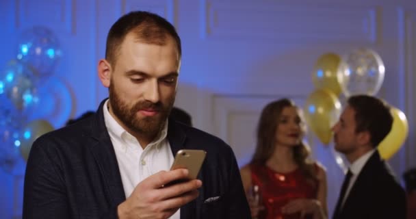 Un joven guapo charlando en su teléfono inteligente y bebiendo champán en la fiesta. Gente bailando y globos detrás. En interiores — Vídeos de Stock