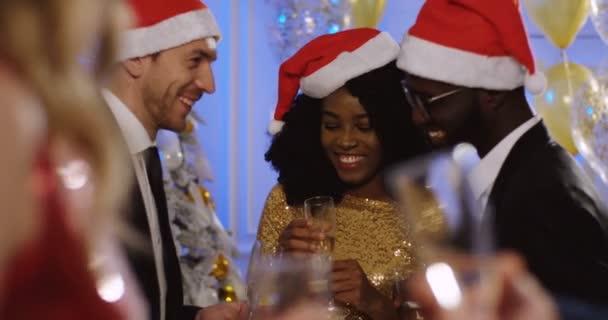 Jóvenes amigos multiétnicos en los sombreros de Santa Claus hablando y bebiendo champán en la fiesta de Navidad. Celebración de Navidad. En interiores — Vídeo de stock