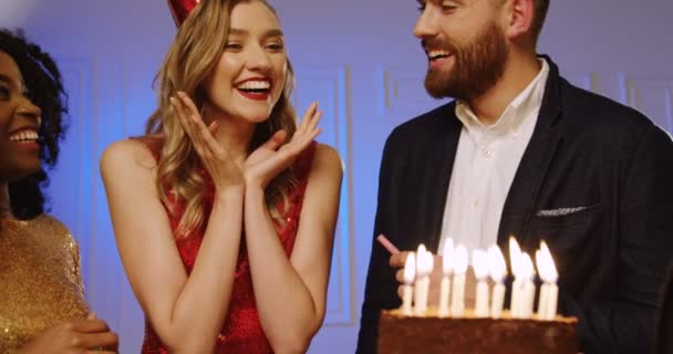 Joven encantadora mujer sorprendente y que soplar velas en un pastel de cumpleaños entre sus amigos en la fiesta. Un retrato. De cerca. De interior — Vídeo de stock