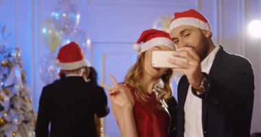 Noel Baba genç mutlu çift portre yapımı güzel selfies yeni yıl partisinde bir akıllı telefon üzerinde şapka. İnsanlar arka dans. Kapalı