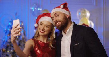 Noel Baba yan tümce şapkaları yılbaşı partisinde bir akıllı telefon komik selfies yapma genç çekici çift portre vurdu. İnsanlar arka dans. Kapalı