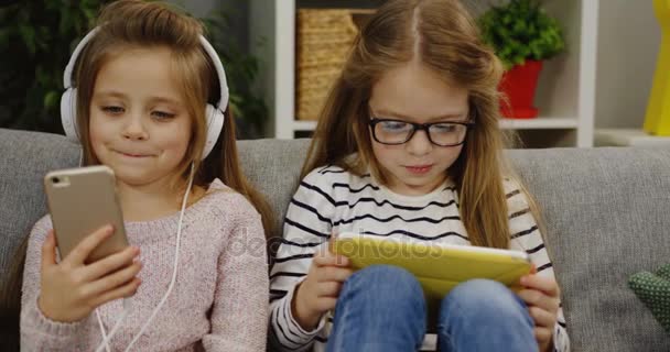 Duas meninas pequenas bonitos sentados no sofá na sala de estar. Uma garota nos grandes fones de ouvido ouvindo a música no smartphone e outra usando um dispositivo tablet. Em casa. Interior — Vídeo de Stock