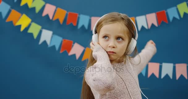 Милая маленькая девочка в наушниках расслабляется и танцует, слушая музыку на синей стене с декорациями фона. Внутрь. Портрет — стоковое видео