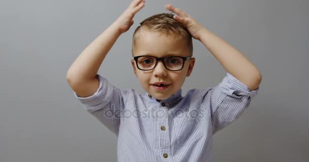 Divertido niñito sin diente y con las gafas sosteniendo la cabeza con los brazos y mirando a la cámara sobre el fondo gris de la pared. De cerca. Retrato. En interiores — Vídeos de Stock