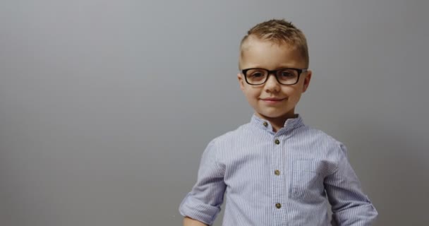 Портрет милого маленького мальчика в очках, показывающего большие пальцы и смотрящего прямо в камеру. на сером фоне стен в помещении — стоковое видео