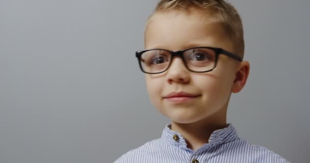 Close up of the lively small blonde boy in the glasses turning his head and looking into the camera with a smile. wall background. Portrait. inside — Stock Video