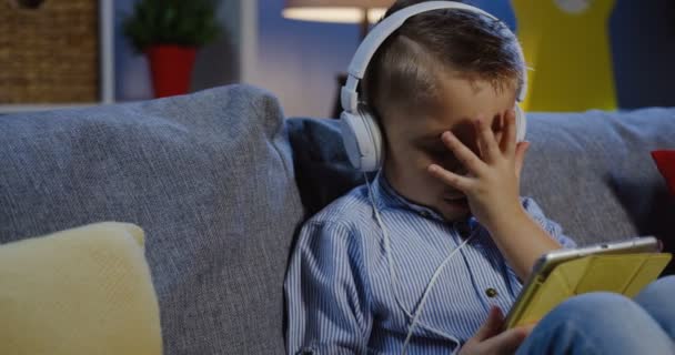 Κοντινό πλάνο της το μικρό ξανθό αγόρι στα ακουστικά κάθεται στον καναπέ στο σαλόνι, γκρι και βλέποντας κάτι στον υπολογιστή tablet, από ό, τι κλείνοντας τα μάτια με το χέρι. Σε εσωτερικούς χώρους. Κατακόρυφος βολή — Αρχείο Βίντεο