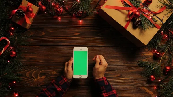 Top view. Woman tapping, scrolling and zooming on the mobile phone vertically. The decorated wooden desk with garlands. Christmas stuff. Green screen, chroma key.