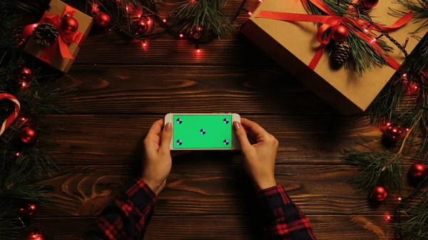 Top view. Woman tapping, scrolling and zooming on the mobile phone horizontally. The decorated wooden desk with Christmas lights. Green screen, chroma key. Tracking motion.
