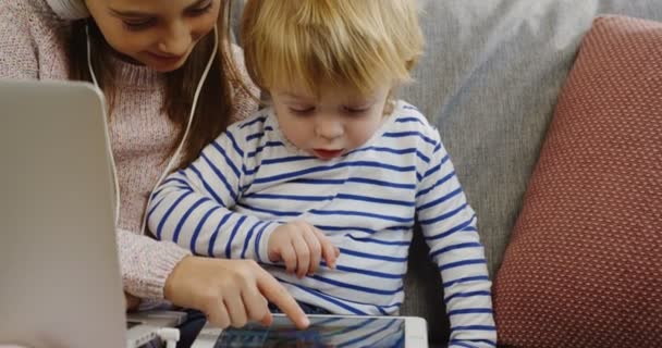 Chica adolescente en auriculares con portátil sentado junto a su hermano pequeño en el sofá y mostrándole cómo jugar juegos en el dispositivo de la tableta. De cerca. De interior — Vídeo de stock