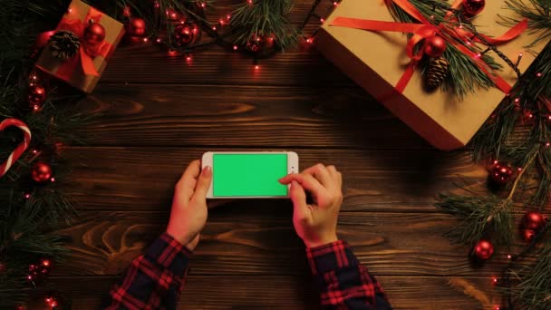 Vue de dessus. Les mains de la femme tapotant, défilant et zoomant sur le téléphone mobile horizontalement. Le bureau en bois décoré avec des lumières de Noël. Écran vert, clé chromatique . — Video