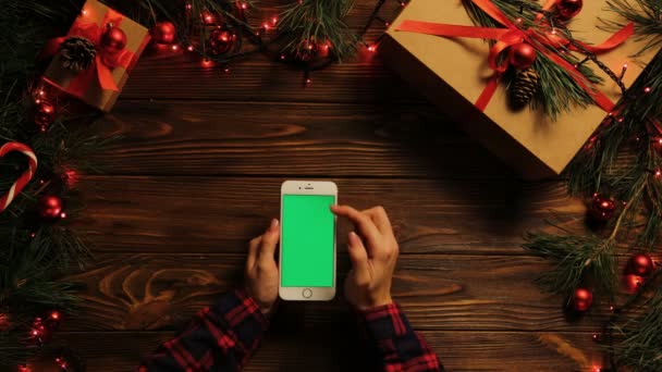 Vue de dessus. Femme tapotant, faisant défiler et zoomant sur le téléphone mobile verticalement. Le bureau en bois décoré avec des guirlandes. Des trucs de Noël. Écran vert, clé chromatique . — Video