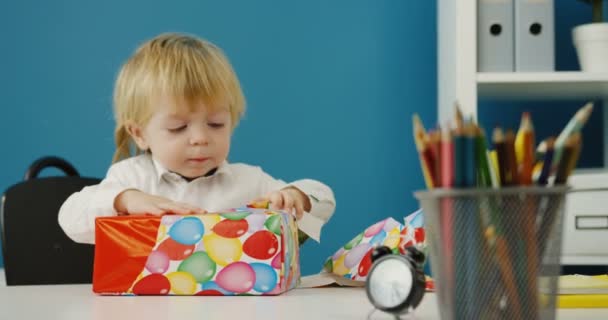 Retrato del lindo niño de camisa blanca desenvolviendo un regalo en la mesa de la oficina. En interiores — Vídeos de Stock