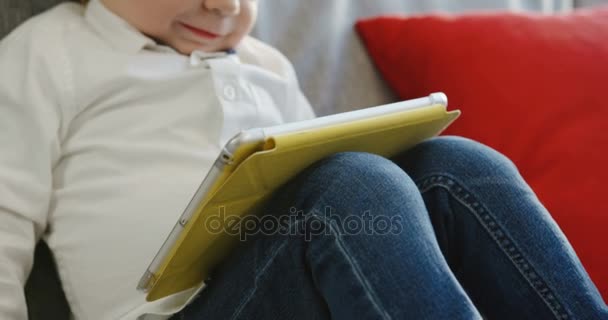 Primer plano del lindo niño pequeño sosteniendo una tableta en sus rodillas y mirando a la pantalla. En casa. Un retrato. De interior — Vídeos de Stock