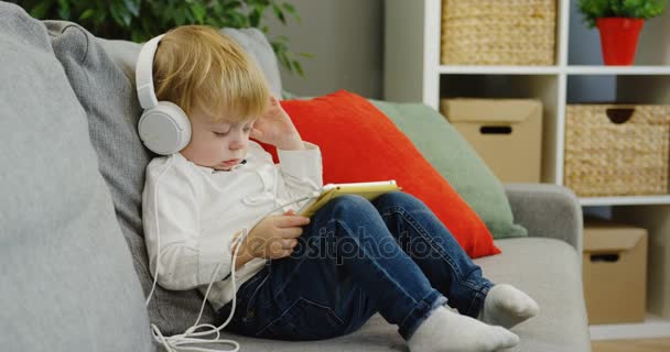 Милий маленький хлопчик у великих білих навушниках слухає музику на планшетному пристрої у затишній кімнаті. всередині — стокове відео