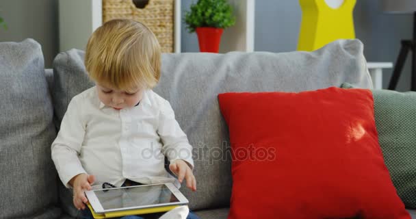 Retrato de la linda rubia niño sentado en el sofá con almohadas y ver algo en el dispositivo de la tableta en la habitación ecozy th. En interiores — Vídeos de Stock