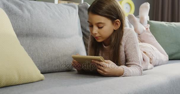 Lovely schoolgirl writing with a pencil in the notebook while lying on the grey sofa in the nice living room. Indoor — Stock Video