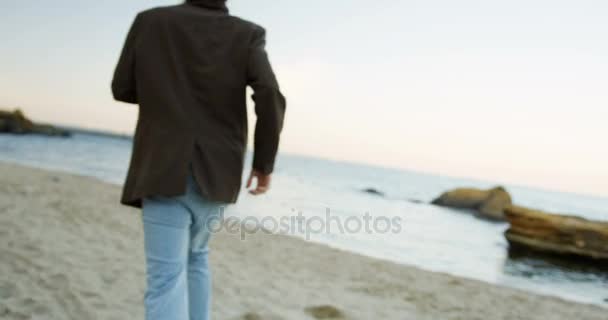 Tir arrière. Gros plan de l'homme en jean et manteau courant sur le littoral. Humeur mélancolique. Temps nuageux. En plein air. Mouvement lent — Video