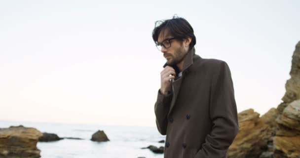 Ein attraktiver Mann mit Brille und Mantel schlendert traurig am felsigen Strand entlang und denkt ernsthaft nach. Draußen. Kaltes Wetter — Stockvideo