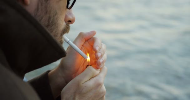 Крупным планом человек в очках с бородой зажигает сигарету на морском фоне. Снаружи. Портретный снимок — стоковое видео