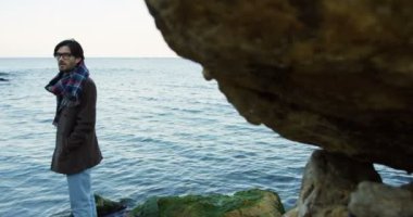 Sahil kayaların üzerinde duran ve kameraya omzunun üzerinden seyir çekici adam. Soğuk hava. Açık havada. Arkadan