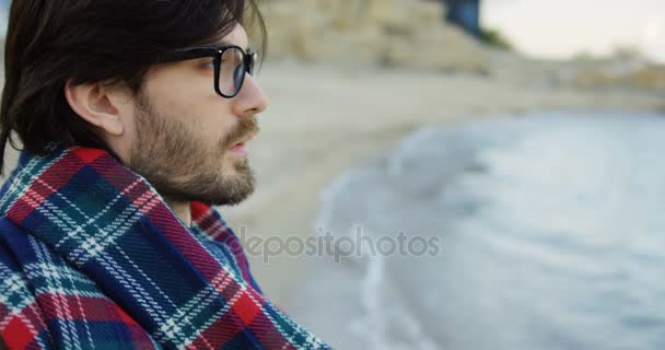 Primo piano di bell'uomo che guarda dritto sulle onde del mare mentre è seduto sulla spiaggia rocciosa. All'aperto. Ritratto — Video Stock