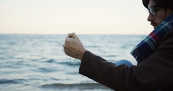 O homem caucasiano bonito sentado perto do mar e polvilhando uma areia em humor deprimente. Dia de tempo nublado. Fecha por trás. Fora — Vídeo de Stock